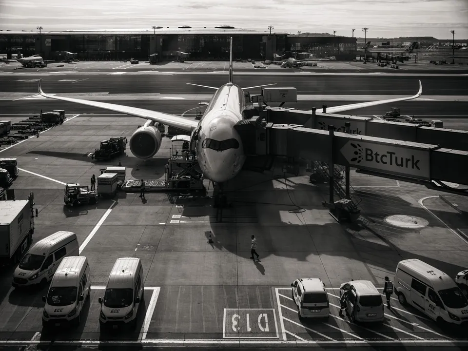 Vista nocturna en blanco y negro de un avión de fuselaje ancho en la pasarela de embarque de una terminal, con vehículos de rampa alineados en primer plano.