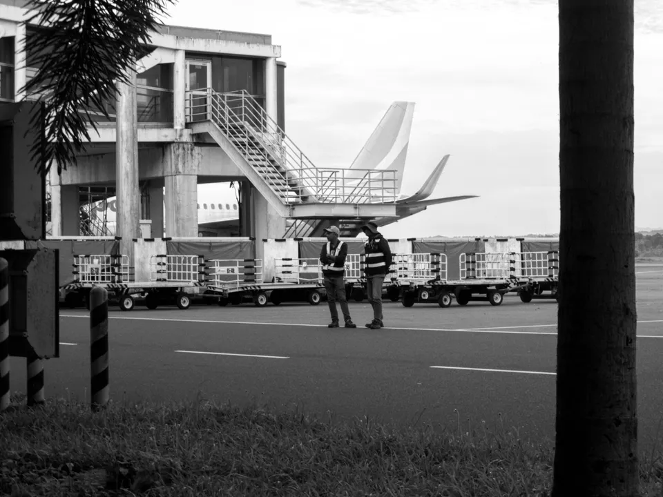 Dos operarios de rampa conversan junto a una fila de carretillas portacontenedores, con las colas de dos aviones y una pasarela de embarque al fondo, en blanco y negro.